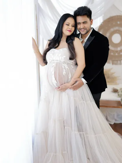 A classic and elegant couple's portrait. The mom-to-be looks radiant in a white gown, standing by the window with her partner.