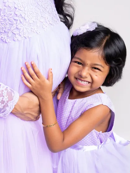 A close-up shot full of personality and joy. The big-sister-to-be gives a huge, happy grin while hugging her mom's belly, showcasing her excitement.