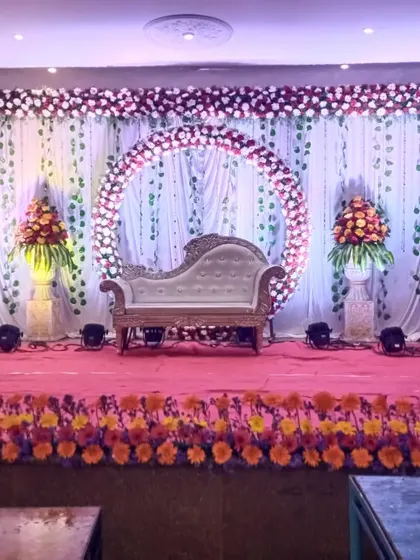 This reception stage combines a circular floral arch with a classic white chaise lounge. The backdrop of white curtains with hanging green vines adds a touch of nature, while the marigold border at the front provides a traditional element.