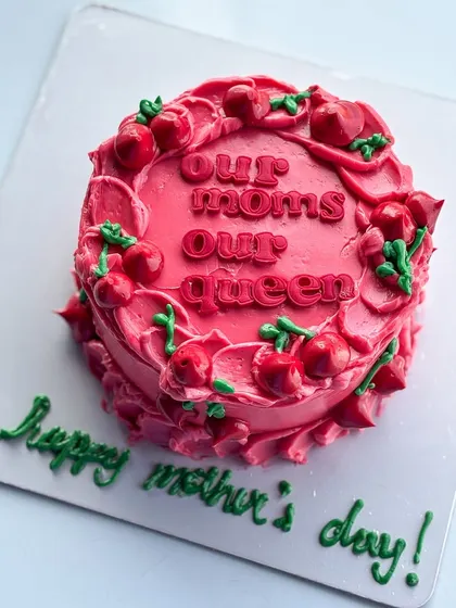 A special cake for Mother's Day with the message "Our Moms Our Queen." The vibrant pink buttercream is decorated with piped cherries for a fun and festive look.