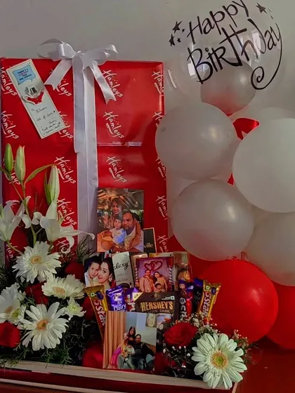 This grand birthday hamper features a large arrangement of fresh white daisies, lilies, and red roses. The flowers beautifully complement the red and white theme of the gifts and balloons.