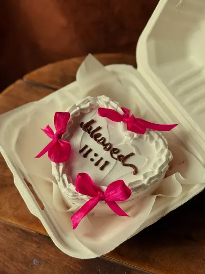 A beautiful heart-shaped bento cake with the word "blessed" and the angel number "11:11" piped on top. The delicate pink satin bows add a sweet, romantic touch.