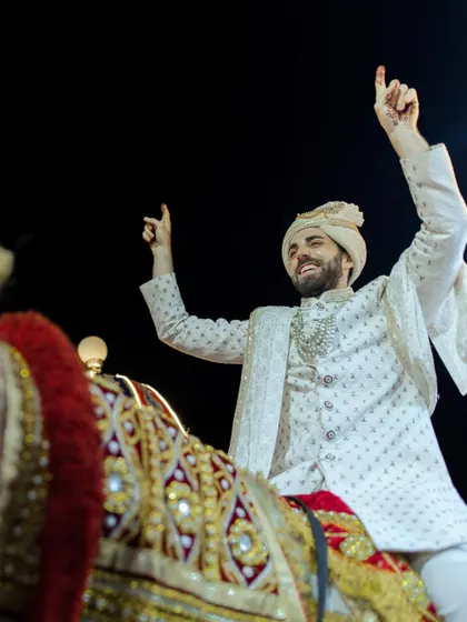 The groom's grand entrance on a decorated horse. His joyful expression and raised hands capture the energy and excitement of a traditional Indian baraat.