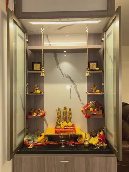 The doors of the pooja unit open to reveal a beautifully organized space with hanging brass bells and ample room for idols and prayer essentials.