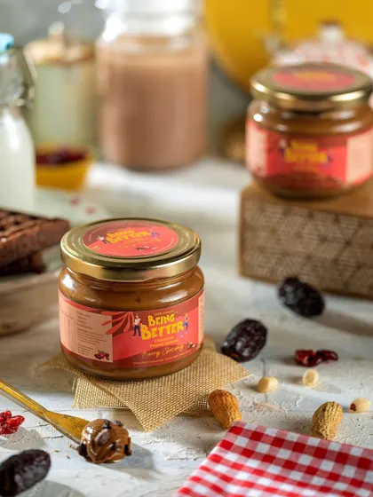 A styled shot of Being Better's peanut spread, with ingredients like dates and nuts scattered around. The composition uses burlap and a checkered napkin to create a rustic, wholesome vibe.