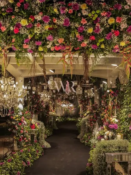 A night view of the floral entrance tunnel. The path is flanked by walls of flowers and greenery, with a chandelier and hanging lanterns adding a warm, inviting glow.