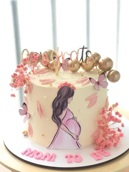 A "Mom to Be" cake with a sweet illustration, pink flowers, and golden balloon details.