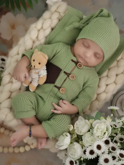 A cozy and cute newborn portrait. The baby is dressed in a sage green knit outfit, cuddling a tiny teddy bear on a miniature bed, creating a heartwarming scene.