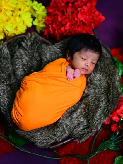 A colorful newborn portrait with the baby in an orange swaddle, nestled in a heart-shaped basket surrounded by bright flowers.