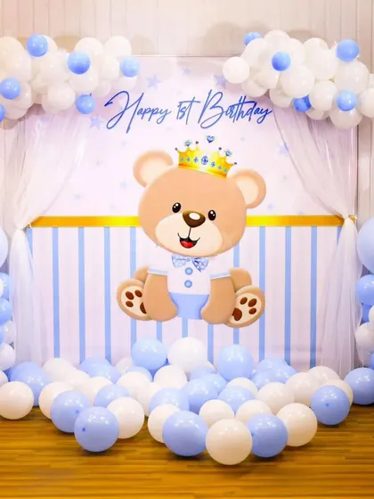 A sweet teddy bear prince theme for a first birthday. This setup features a custom backdrop with a cute bear, framed by blue and white balloon garlands and columns.