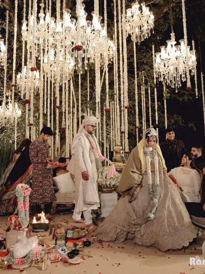A wide shot of the wedding ceremony, showing the couple at the mandap under a canopy of hanging flowers and chandeliers, capturing the beautiful and grand decor.