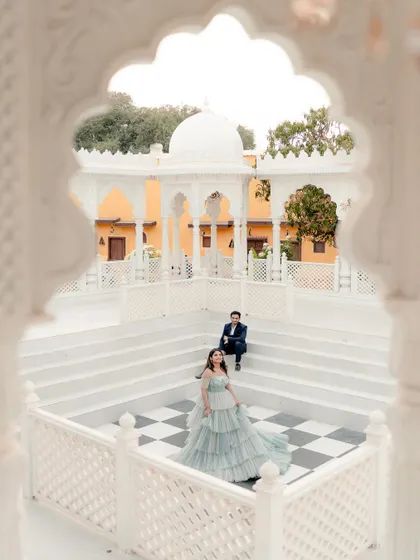 A fairy-tale pre-wedding shot. The couple is framed by a white marble archway, with the bride in a flowing gown, creating a magical scene.