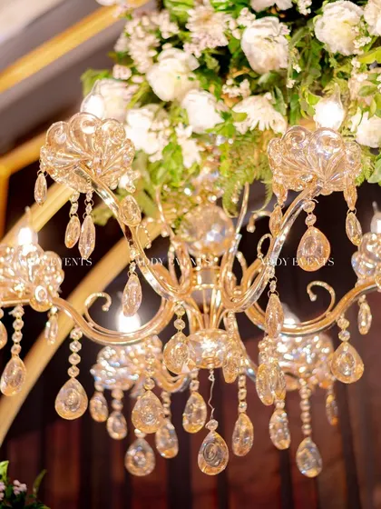 A close-up view of a crystal chandelier nestled within a lush arch of white flowers. This combination of sparkling crystal and soft petals is a signature element of our luxurious and romantic stage designs.
