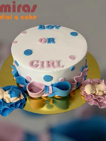 A classic "Boy or Girl?" gender reveal cake. The white cake is decorated with pink and blue polka dots and bows, with tiny sleeping babies in blue and pink on the side.