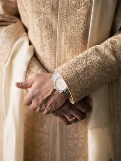 A detail shot of the groom's hands, showing his watch and ring, against the rich texture of his embroidered sherwani.