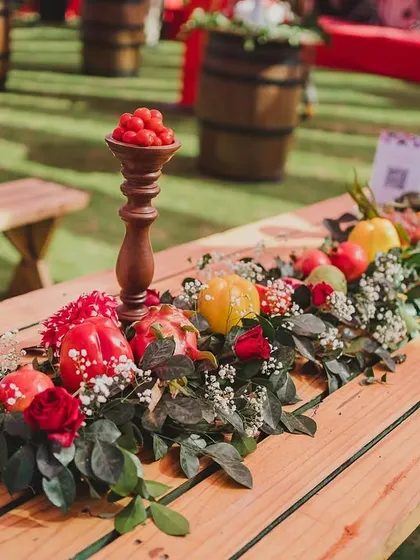 The tables were designed as rustic picnic benches with runners made of fresh vegetables, fruits, and flowers. This centerpiece features red roses, baby's breath, bell peppers, and pomegranates on wooden candle stands.