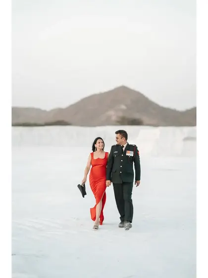 A wide shot of the couple walking hand-in-hand in a surreal white landscape, the groom in his uniform and the bride in a striking red dress.