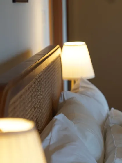 The warm glow from a simple bedside table lamp reflects softly on a rattan headboard. This image captures the serene and restful feeling I aim to create with my designs.