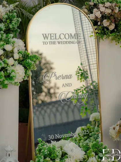 A personalized welcome mirror, nestled amongst soft white and green florals, greets guests with elegance and warmth.