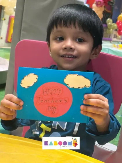 A young boy happily shows the "Happy Teacher's Day" card he made, decorated with a bright sun and fluffy clouds. Simple, heartfelt, and made with love.