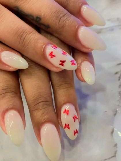 A delicate and pretty design on milky white almond nails. The ring fingers are accented with tiny, hand-painted red butterflies for a touch of whimsy.