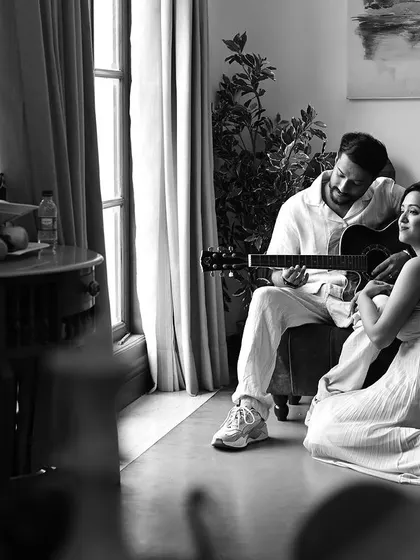 A black and white indoor shot creating a serene, musical moment. The composition, looking past a foreground object, adds a cinematic, observational quality to the image.