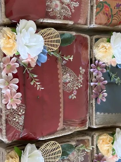 Close-up of a deep red saree packed in a jute basket. The intricate embroidery of the saree is complemented by the delicate floral arrangement on top.