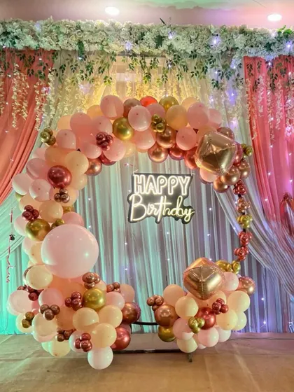 A glamorous birthday photo booth with a circular balloon arch in shades of pink, peach, and gold. The backdrop of drapes and a neon sign adds to the festive look.