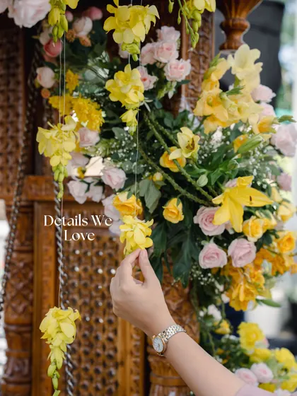 A team member carefully places a hanging floral element, showcasing the hands-on attention to detail that goes into every setup. Here, bright yellow flowers are paired with a traditionally carved wooden structure.