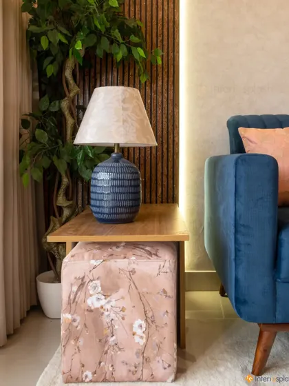 A detail shot of a cozy living room corner. The blue table lamp complements the sofa, and the floral-patterned ottoman adds a soft, decorative touch.