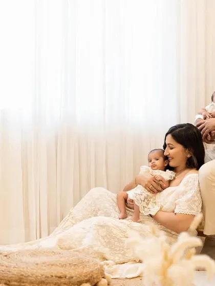 A different perspective of the new family of four. The mother sits on the floor holding one twin, while the father sits on a bench behind her with the other, creating a layered and interesting family composition.