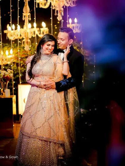 A romantic portrait of a couple at their engagement party. The groom holds his bride close as they share a look, with beautiful chandelier lights creating a magical bokeh effect.