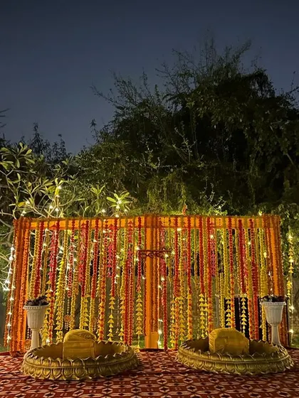 An outdoor Haldi setup with a backdrop of hanging marigold strings. The low seating arrangement with large brass platters is perfect for a traditional ceremony.