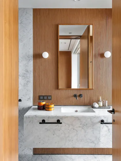 The adjoining bathroom continues the bedroom's language with minimal white Carrara marble and fluted wood accents. This visual continuity ensures a seamless transition between the sleeping and bathing areas.