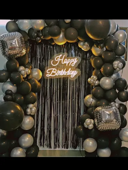A bold black and silver birthday setup. This balloon arch frames a black shimmer curtain and is lit by a 'Happy Birthday' neon sign.