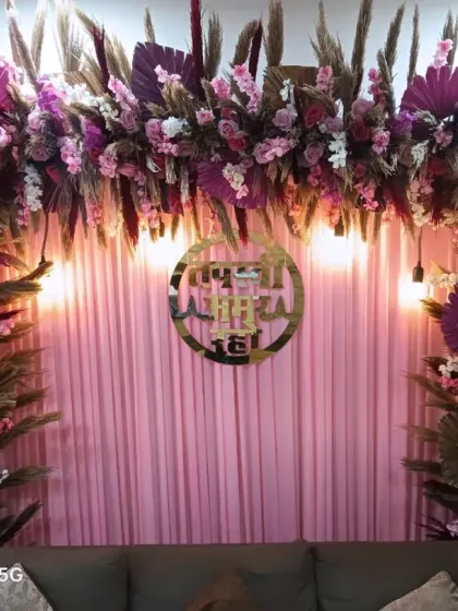 A dense floral arch with pink and purple flowers mixed with pampas grass. This backdrop for a Jain ceremony features a golden sign and is set against a pink curtain.
