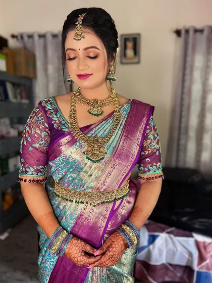 This bride from Mysore looks radiant in a unique peacock-toned silk saree. Her makeup features a soft pink and gold eye look with a matching lip color to complement her intricate floral blouse.