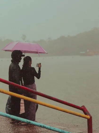 A couple takes a photo with their phone by the lake, sharing a pink umbrella.