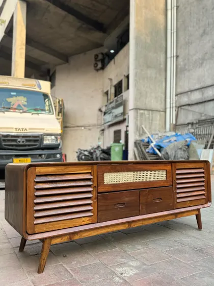 A mid-century modern media console made from solid teak. It features a mix of storage options including slatted doors, drawers, and a central open shelf with a cane back.