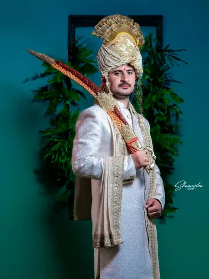 A groom in a majestic white sherwani and golden turban, holding a ceremonial sword. The indoor lighting and simple background put all the focus on his regal attire.
