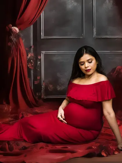 A serene, seated portrait from a fine art maternity session. The setup includes a floor covered in red fabric and petals, with dramatic curtains and florals in the background, creating a rich and romantic atmosphere.