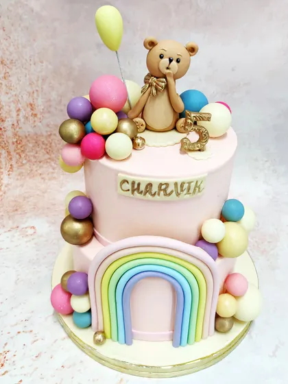 We combine several whimsical themes in this two-tiered rainbow teddy bear cake. A sweet teddy bear holding a balloon sits atop a pink cake adorned with a pastel rainbow and a cascade of colorful spheres, creating a joyful and imaginative design.