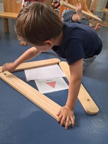 A young boy constructs a triangle using wooden planks, guided by a reference card. This hands-on approach makes learning about geometry intuitive and fun.