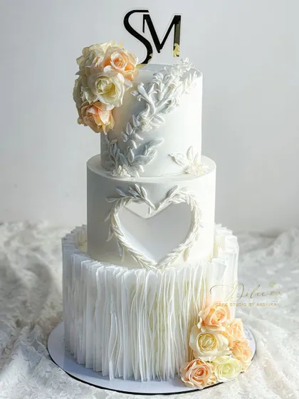 A wedding cake should be as beautiful as the love story it celebrates. This all-white, three-tiered fondant cake features delicate wafer paper ruffles, a heart-shaped cutout, and intricate leaf patterns. The soft peach and cream sugar flowers add a touch of timeless elegance.