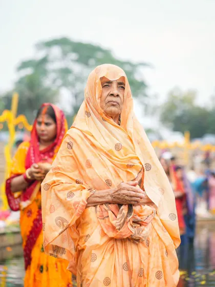 An elderly woman stands with folded hands, her face reflecting a lifetime of faith and devotion during Chhath Puja. The festival brings together people of all ages.