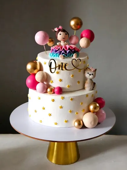 A sweet and simple first birthday cake. This design features a girl figurine with her cat, surrounded by pink, gold, and white balloons.