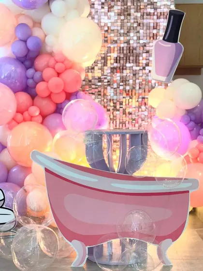 A focused shot of the spa party's central props, the pink bathtub and a nail polish cutout. The background features the shimmering sequin wall and a cascade of colorful balloons, emphasizing the fun, thematic elements.