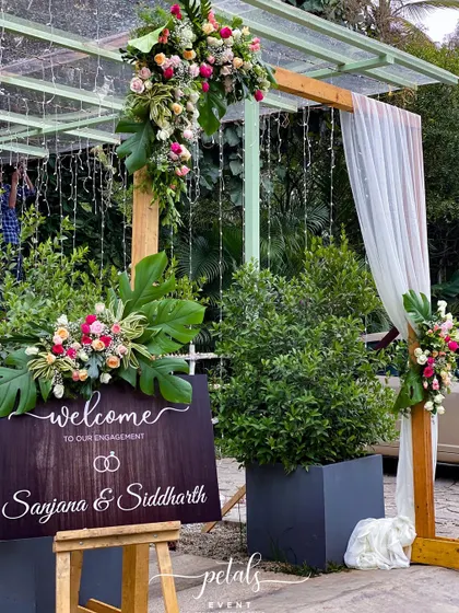 A welcoming entrance for an engagement party, featuring a wooden arch, soft drapes, and a personalized sign. The arrangements of pastel roses and tropical leaves add a touch of romance.