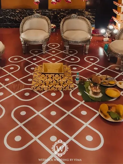 A traditional red and white mandap setup inside a grand hall. The decor features pillars adorned with diyas and a ceiling covered in hanging garlands of jasmine and red roses.
