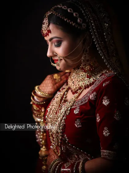 A classic bridal portrait against a dark background. The lighting perfectly highlights the rich details and colors of her velvet lehenga and gold jewelry.